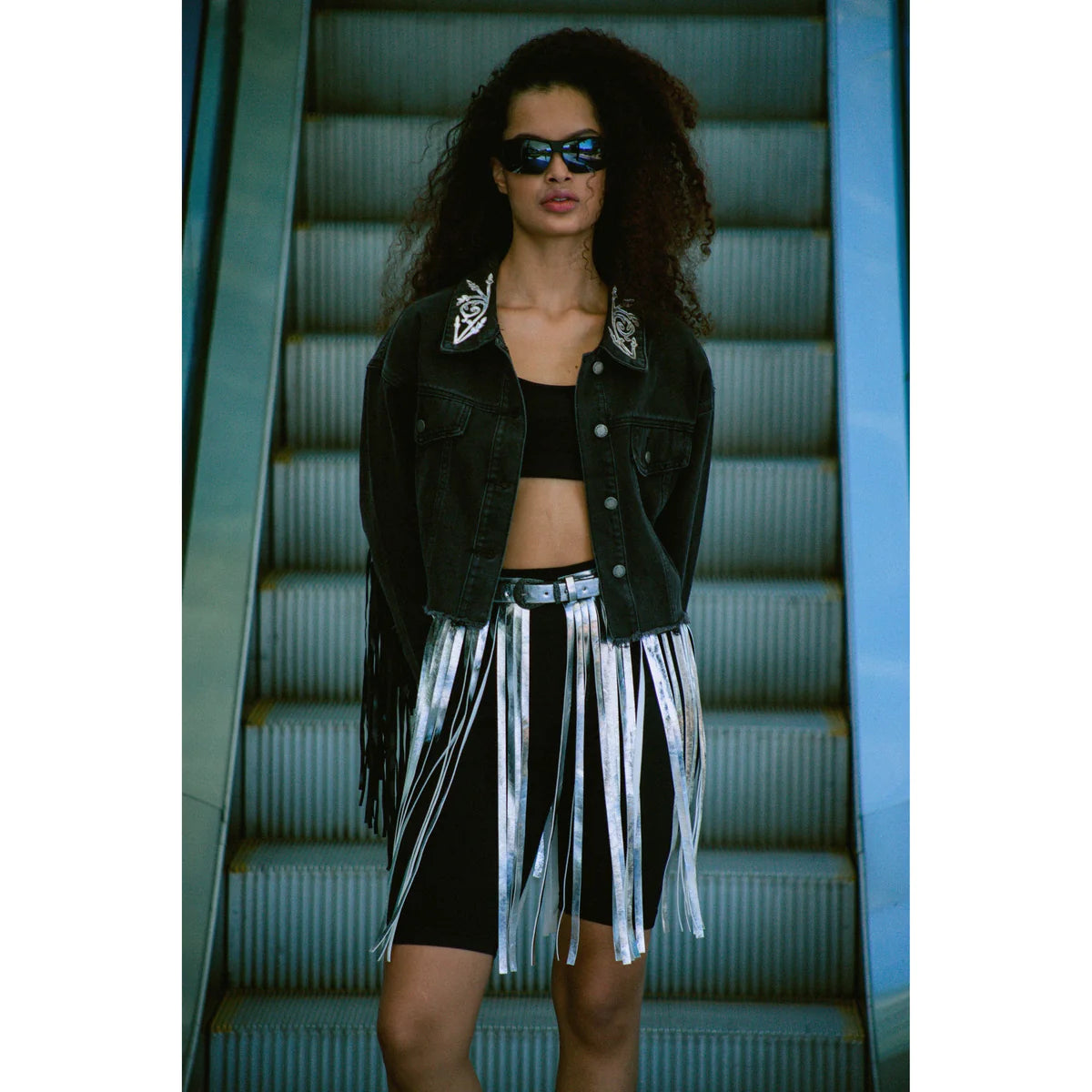 a curly hair model showing us how to style sunglasses with an electric stair as a background a black top and a leggins denim rag skirt by OnPost